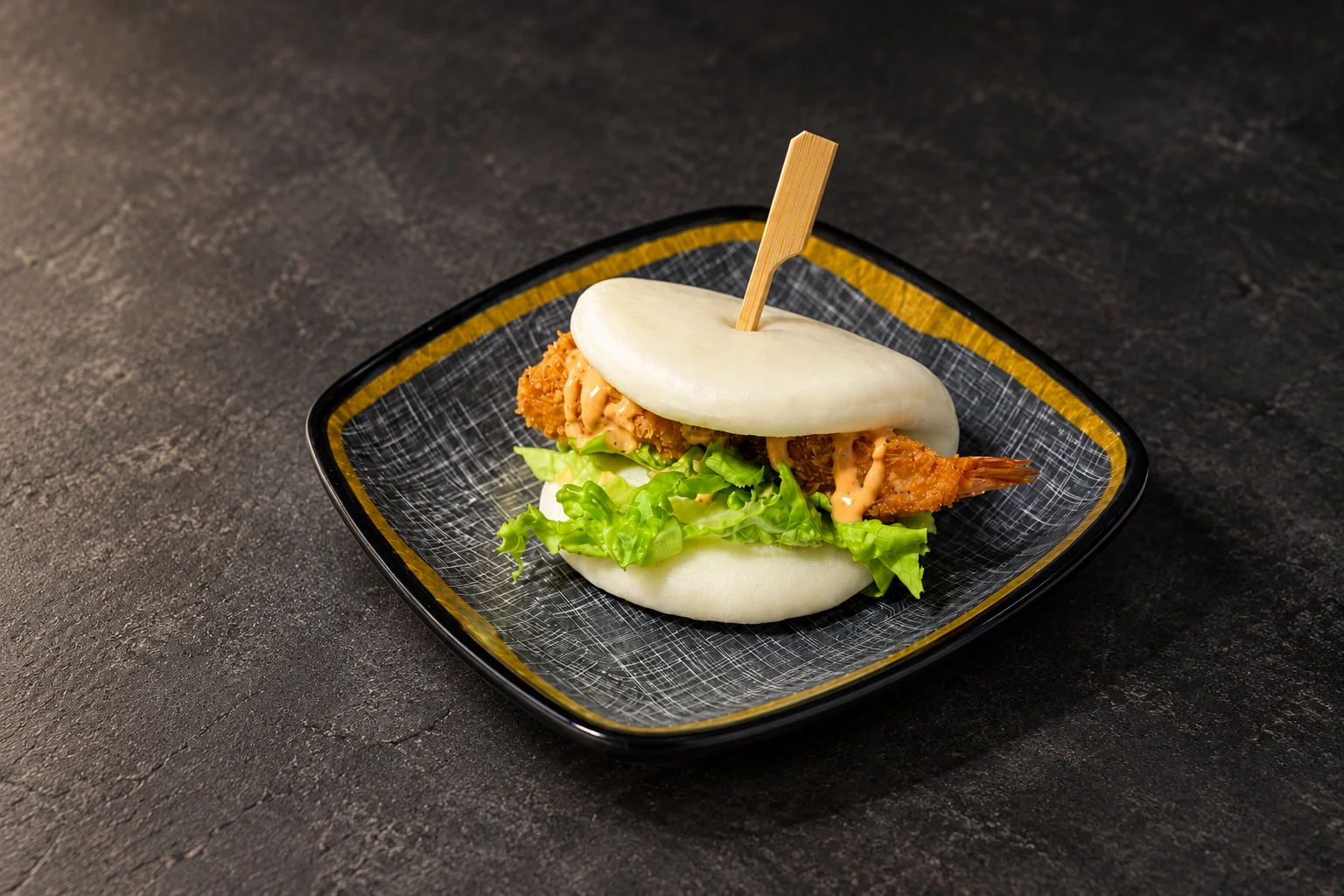 Pain bao garni d'une crevette tempura et de salade, sur une assiette carrée à liseré doré.
